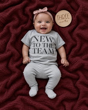 Baby on a burgundy blanket wearing a naturally draped NEW TO THE TEAM tee