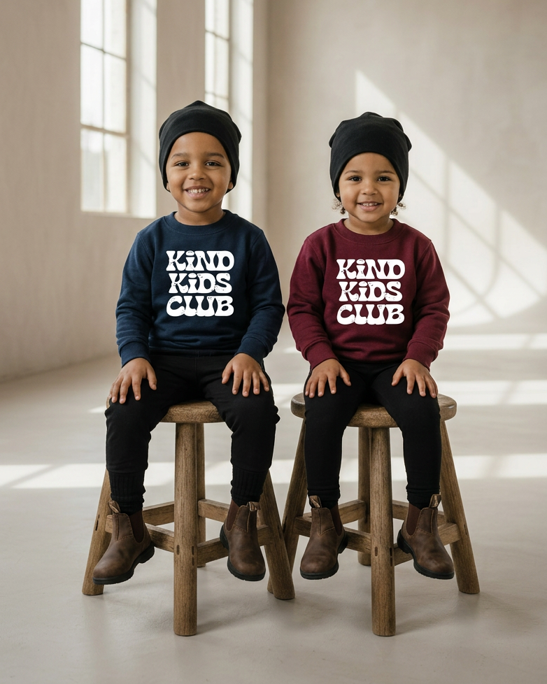 Boy and girl seated together in navy and burgundy Kind Kids Club crewnecks with white lettering, black beanies, and brown Chelsea boots