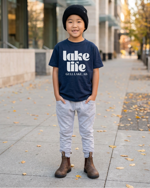 Boy wearing a navy lake life shirt with white lettering, a black beanie, and brown Chelsea boots in a modern fall city setting, with room for a 4:5 crop