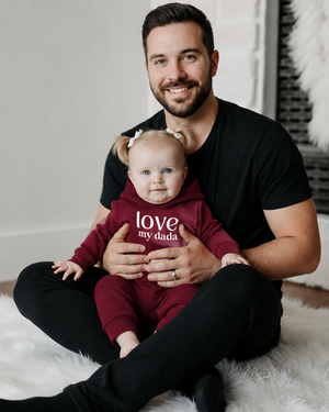 Little girl in a burgundy outfit with white love my dada lettering sitting with her father