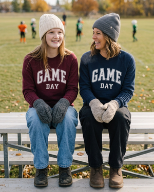 Two women in burgundy and navy Game Day crewnecks watching youth football in autumn